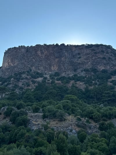 The image shows the cliff face of a mountain near Seydikemer, Turkey, adorned with numerous ancient rock-cut tombs. These tombs, resembling small openings or windows, are intricately carved into the sheer rock surface, creating a striking visual. The mountain is covered in dense green vegetation, with trees and shrubs growing on its slopes and along its base. The sky above is a clear, pale blue, with the sun partially obscured by the crest of the mountain, casting a subtle glow. The scene evokes a sense of history and natural grandeur.
