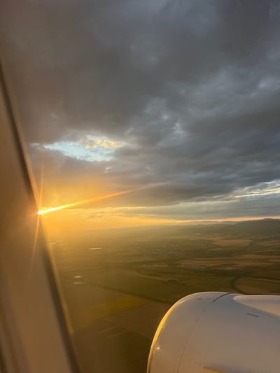 An aerial view from an airplane window captures a sunset over a rural landscape. The sun, a bright orb, is low on the horizon and partially obscured by clouds, casting a golden light across the scene. The sky is a mix of dark, dramatic clouds and patches of lighter, illuminated sky. Below, a patchwork of agricultural fields stretches towards distant hills. The wing of the airplane is visible in the foreground, a smooth, white surface with the words "NO STEP" printed on it. The location is identified as Grigorevo, Bulgaria.