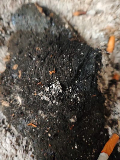 The image shows a close-up of an ashtray containing cigarette butts and ash. The ashtray is filled with gray and black ash, and several orange cigarette butts are visible. The surface has a rough texture with small particles and debris scattered around. There is no indication of the specific location in Aumetz, France, or any other details about the environment, people, or actions.