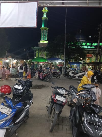 This night-time image depicts an active public square or market area in Sumberejo, Indonesia, adjacent to a mosque. In the background, a tall minaret, brightly illuminated with green lights, rises above a mosque building that also features green illumination and displays the text "CENTER AL AMAN PEMBANG."

The midground is densely populated with individuals engaged in various activities around numerous illuminated food stalls. Several people are visible standing and sitting near these stalls, some of which are covered by green and blue umbrellas. Parked motorcycles are interspersed among the people and stalls, including a white Honda PCX scooter.

In the foreground, several motorcycles are parked on a paved surface composed of paver blocks. From left to right: a blue and white Honda scooter (license plate K 6845 DW, expiration 04-28) with a red helmet resting on its handlebars; a black scooter (license plate K 2003 WO, expiration 08-23); a red and black Honda motorcycle (license plate K 5419 HM, expiration 11-27); and a black scooter (license plate K 6084 MW, expiration 03-24) with a black and orange helmet on its seat. A woman wearing a yellow headscarf and top is positioned near the rightmost black scooter, seemingly interacting with it, while a small child in light-colored clothing stands beside her.

At the top of the frame, a large, blank white rectangular banner or screen is suspended horizontally. The scene is artificially lit, creating distinct areas of brightness around the stalls and mosque against the dark night sky.