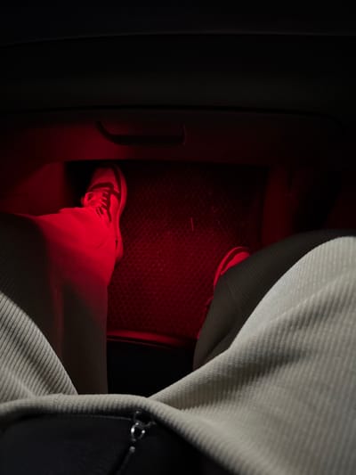 A close-up shot, heavily tinted in red light, reveals a person's foot wearing a white sneaker in a car's footwell. The floor mat has a textured surface. The person is wearing light-colored corduroy pants. Part of the center console is visible on the left side. A bag or jacket with a zipper is also visible on the right side. The overall scene is dimly lit and slightly distorted by the red hue.