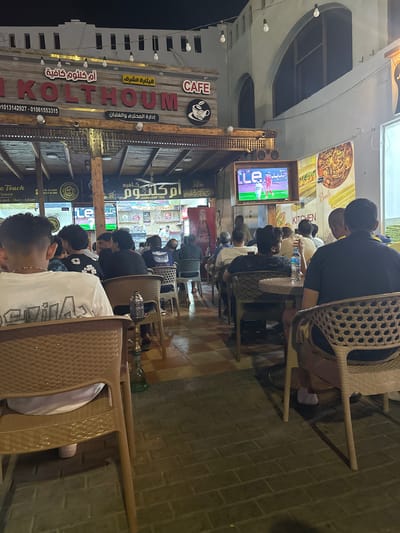 The scene depicts an outdoor cafe, potentially in Dahab, Egypt, given the Arabic signage ("Om Kolthoum Cafe") and the overall ambiance. The cafe is bustling with patrons, mostly men, seated on wicker chairs and tables. They appear to be watching a soccer game on a television screen mounted higher up in the area, the broadcast clearly visible. Shisha pipes are present on some tables, suggesting a relaxed social setting. The cafe has a rustic aesthetic with wood accents and signage in both Arabic and English. Coca-Cola branding is visible, indicating the drinks served. The time of day appears to be evening or night, with artificial lighting illuminating the scene. A brick paved road is visible.