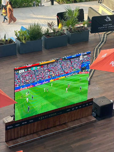 The image shows an outdoor scene at Marina Puerto Cancún, Mexico, on a wooden deck. A large outdoor LED screen displays a live soccer match between Morocco and Scotland, with the score currently 0-1. The match is in the 76th minute and 25 seconds. Spectators are visible in the background on the screen. In the foreground, several people are walking past the screen, including a woman in a long patterned dress and a child in a yellow shirt. Various potted plants are arranged in grey rectangular planters. An orange canopy or umbrella is partially visible in the lower right corner. A black speaker box is positioned to the right of the screen. The overall environment suggests an open-air entertainment or viewing area within the marina complex.