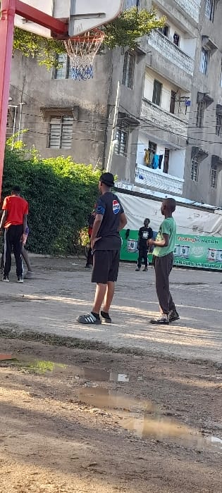 This image depicts an outdoor scene in Frere Town ward, Kenya, featuring multiple individuals and urban structures. In the foreground, an unpaved dirt surface shows several puddles reflecting the sky and surrounding elements.
To the left, a red-painted metal basketball hoop structure with a white net and blue accents is visible. Behind the hoop, a dense green hedge or bush spans the midground.
In the midground, five male individuals are present. The most prominent figure, facing away from the camera, wears a black t-shirt with a blue and red Pepsi logo on the back, black shorts, and black Adidas slide sandals over white socks. He stands with his hands on his hips. To his right, another male, facing left, wears a light green t-shirt and dark pants, appearing to observe. Further left, a third male in a red t-shirt and dark pants stands with his back partially to the camera. Two additional figures are partially visible in the background; one near a white and green stall, wearing a dark t-shirt.
The background features a multi-story concrete building with numerous windows and balconies. Clothes are visible hanging on a line from one of the lower balconies. To the right of the central figures, a white tent-like structure or vendor stall with green trim displays text including "Feeding the NATION" and "MAHOUD'S SHAWARMA," along with images of white containers.
The scene is illuminated by natural light, casting subtle shadows and suggesting daytime conditions.