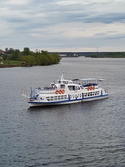 The image depicts a white passenger riverboat with blue trim and orange life preservers on a body of water, presumably the Volga River. The boat is traveling on the water away from the shore in the direction the boat is pointed. On the shore are trees and a few indistinct buildings, and people on the lawn. Further in the background is a thin bridge or tower. The sky is overcast. It is daytime. The location is Faliukovo, Russia.
