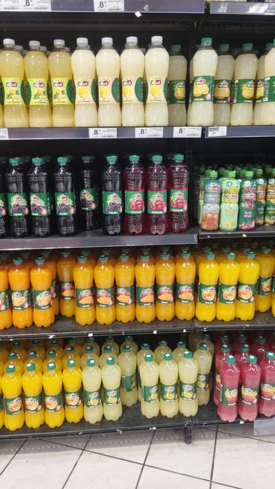 The image shows a shelf in a supermarket in Kfar Saba, Israel, stocked with various bottled beverages. The products are arranged in rows on metal shelves. On the top shelf, there are bottles of light-colored beverages, likely juices or flavored water, with labels featuring lemons and other fruits. Below that, there are rows of dark purple and red bottled beverages, which appear to be grape and berry juices. The bottom shelf is filled with orange and yellow bottled drinks, predominantly mango and possibly other tropical fruit juices. Some bottles have green caps, while others have white or yellow caps. The labels are in Hebrew. The environment is a retail setting, with the typical appearance of supermarket shelving. There are no people or animals visible in the image.