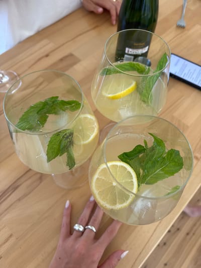 Three large, round glasses of a pale yellow beverage garnished with lemon slices and fresh mint sprigs rest on a light-colored wooden surface. A green bottle partially visible near one glass suggests sparkling wine or champagne as an ingredient. A smartphone and a fork are also in the background. A hand adorned with two rings rests near one of the glasses, adding a personal touch to the scene.