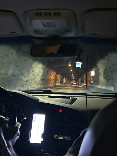 This media file shows the interior of a vehicle driving through a tunnel at night. The driver's hand is visible on the steering wheel, and a smartphone mounted on the dashboard displays a navigation app. The tunnel appears to be narrow, with walls lined with lights. The road surface is wet and reflective, suggesting recent rain or melting snow. There is a dashcam mounted on the windshield, recording the journey. A rear-view mirror is also visible, reflecting the tunnel ahead. The lighting in the tunnel is a mix of artificial lights and the headlights of the vehicle. The overall mood is one of focused travel through a dark and enclosed space. Text visible on the dashboard reads "ERROR". The image's orientation suggests it was taken while moving.