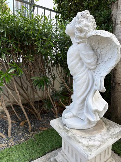 A white plaster statue of an angel stands on a pedestal in a garden. The statue is positioned in the foreground, facing away from the viewer and looking upwards. The angel has feathered wings, curly hair, and is draped in flowing robes. The pedestal is weathered and shows signs of age. Behind the statue, there is a dense shrub with green leaves and thin branches, and a portion of a tree trunk is visible on the right side of the frame. The ground is covered with small dark stones and a strip of green grass. A metal fence can be seen in the background. The setting appears to be an outdoor residential area in Bang Kaeo Subdistrict, Thailand, given the vegetation and the style of the statue and fence, commonly found in Thai gardens.