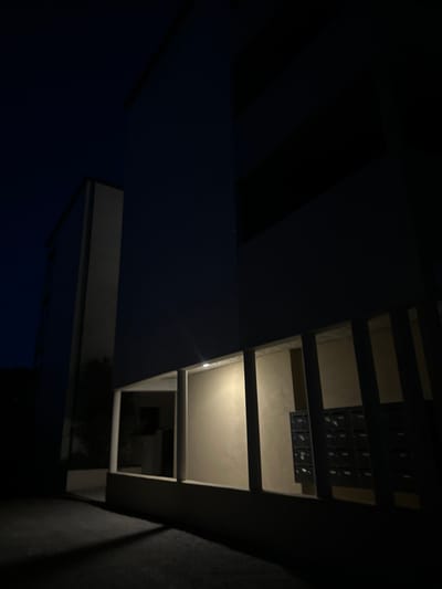 The image depicts the exterior of a modern building at night, likely in the city of Anduze, France. A tall, rectangular structure with a white facade is visible on the left. To the right, a darker building section recedes into the darkness. In the foreground, a walkway or patio area is illuminated by soft light emanating from within a sheltered section of the building. This sheltered area is characterized by a light-colored wall and a series of vertical support beams. Mounted on the wall are rows of dark, compartmentalized mailboxes, suggesting this is an entrance or common area. The ground in front of the building is dark and appears to be asphalt or concrete, with shadows cast from the structure. The sky above is a deep, dark blue, indicative of nighttime. There are no people visible in the scene.