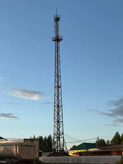 A tall, lattice-style telecommunications tower stands centrally in the frame, dominating the view against a clear blue sky with scattered wispy clouds. The tower is dark metal and appears to be a significant height, with multiple levels and platforms visible. In the foreground, the upper part of a large, light-colored truck, possibly a dump truck, is visible on the left side of the image. Behind the truck and to the right of the tower, several low-rise buildings with wooden siding and darker roofs are situated. Trees are visible behind the buildings, suggesting a semi-rural or suburban environment. A banner with Russian text is stretched across the front of one of the buildings. The lighting suggests it is late afternoon or early evening, with the sun casting a warm glow on some of the structures. The image captures a scene in Staraya, Russia.