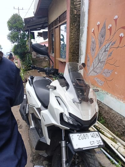 The image displays a white Honda ADV 150 scooter parked outdoors on an unpaved surface. The scooter's front license plate, "F 2089 HBN" with "07-20" below, indicates registration consistent with Indonesia, specifically the West Java region, rather than Pinnwe, Myanmar (Burma). The scooter features a clear windshield, black handlebars, and integrated turn signals.

To the right of the scooter, an orange wall is visible, decorated with a hand-painted mural of stylized grey leaves and orange flowers with white tips. Below this mural, several white pipes, partially covered in green growth, lie against the wall. A rough-textured vertical concrete pillar stands to the right of the scooter, separating the orange wall from an adjacent building with a beige wall and wooden-framed windows.

In the immediate foreground on the left, the back and right shoulder of an individual wearing a dark blue short-sleeved shirt are partially visible. In the background, an unpaved path ascends, along which a blurry figure, possibly a woman in light-colored clothing, walks away from the viewer. Green foliage and small trees line this path. Behind the foliage, a building with a corrugated metal roof overhang is present, from which clothes are hanging to dry. Utility poles and wires are visible against an overcast sky in the upper left quadrant of the image.