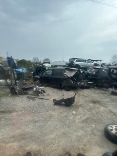The image depicts an outdoor automotive salvage yard located in Hrishteni, Bulgaria, under an overcast sky. The ground consists of light grey gravel mixed with dirt, showing evidence of vehicle and foot traffic.
In the midground, a black multi-purpose vehicle (MPV), possibly a Volkswagen Touran, is prominently displayed in a state of advanced disassembly. Its rear hatch, bumper, and portions of its side panels are absent, exposing internal structural components. The vehicle rests directly on the ground, missing all wheels. To its right, a dark-colored sport utility vehicle (SUV), possibly a Nissan Qashqai/Rogue, is visible, also partially disassembled with its rear hatch removed and internal components exposed. Another partially disassembled dark-colored vehicle is adjacent to the SUV. Behind these, a silver station wagon, possibly a Volkswagen Passat, is stacked atop other vehicle wreckage. Numerous other vehicle bodies and frames are piled in the background, forming distinct mounds of scrap metal.
In the foreground and lower-midground, various vehicle components are scattered across the gravel. These include a black plastic underbody shield, suspension parts, and other metallic fragments. A single vehicle tire with a silver alloy rim is visible in the lower-right corner.
On the left side of the image, a chain-link fence with green metal posts separates a section of the yard. Behind this fence, a blue truck cab or heavy vehicle component is visible, along with stacks of tires and other miscellaneous items. Further back, bare-branched trees and a light-colored building or storage unit are visible against the horizon. No individuals are present in the scene.