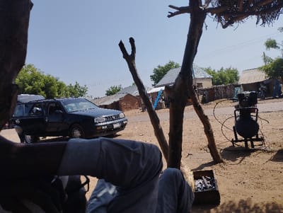 The image was taken in Tongo, Nigeria (10.664, 11.364). It shows a partially obscured view of a person sitting under a large tree in a dusty, dirt-road setting. The person's legs are visible, clad in dark pants and a dark shirt. The tree's branches stretch out, partially obscuring the view of the surroundings. A black car is parked in the foreground on the road. The car is a Volkswagen Golf, and it's facing away from the camera. A metal object, potentially a compressor, is sitting on the ground to the right of the car. In the background, there are a few structures and buildings that appear to be in a residential area. There are trees on the left side of the image and some palm trees in the background. The image appears to have been taken in daylight on a clear day.