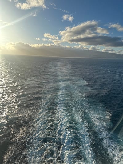 The image depicts an expansive view of the open sea with a distant landmass under a partially cloudy sky. The perspective is from the stern of a moving vessel, evidenced by a prominent, frothy white and light blue wake trailing from the bottom center and extending into the middle distance. Sunlight streams from the upper left, creating a bright glare on the water's surface to the left of the wake, while the water to the right appears a deeper blue. The distant horizon reveals a low mountain range or island, identified as the coast of San Sebastián de La Gomera, Spain, appearing hazy under a band of clouds. The sky is a clear blue, dotted with scattered white cumulus clouds, particularly concentrated above the distant land. No individuals, specific objects beyond the natural elements, or direct interactions are visible within the scene.