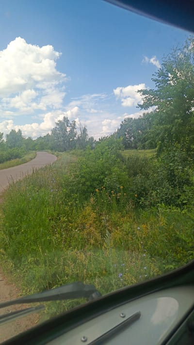 A rural road curves through lush green fields and trees under a partly cloudy blue sky. The view is from inside a vehicle, with a windshield wiper visible in the foreground. The scenery suggests a summer day in a countryside setting, possibly in the Rybushka region of Russia, given the context.
