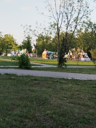 The image depicts a playground with several play structures in Roshchinsky, Russia. In the foreground, a paved pathway curves through a grassy area with a small bush. In the middle ground, a larger playground area is visible, featuring brightly colored structures, including what appears to be a tunnel, slides, and climbing frames. Several children and adults are present on and around the playground equipment. The background is dominated by trees and a clear sky. The overall scene suggests a public park or recreational space on a clear day.