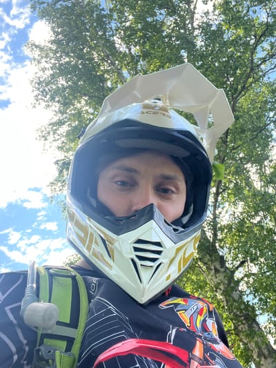 An individual is depicted from a low-angle perspective outdoors in Новочувашский, Russia. The person is wearing a white and gold full-face Acerbis motocross helmet, which obstructs most of their face except for their eyes, nose, and upper cheeks. A small action camera is mounted on the top visor of the helmet. They are wearing a black long-sleeved shirt with a white geometric pattern on the left side and a colorful graphic design featuring red, yellow, orange, and grey elements on the right. A bright green hydration pack with a clear drinking tube and grey bite valve is visible over their left shoulder. The background consists of a blue sky with white cumulus clouds on the left and dense green foliage of deciduous trees on the right, with sunlight visible filtering through the leaves.