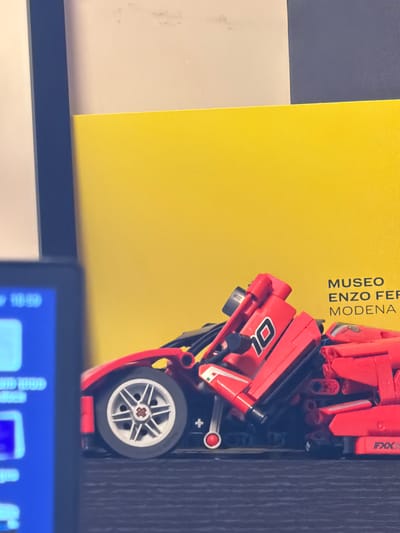 The image displays an indoor, still-life arrangement featuring a red toy car positioned next to a yellow book or display item. The toy car, a Lego-style model, possibly a Ferrari Technic series, has the number "01" clearly visible on its side and the text "FXX K" on a rear spoiler-like element. It features silver multi-spoke wheels with black tires and distinctive red and black building block components.

Adjacent to the car, a bright yellow book or folder stands upright, bearing the black, vertically-oriented text "MUSEO ENZO FERRARI MODENA," indicating its association with the Enzo Ferrari Museum in Modena, Italy. The car itself rests on a dark, textured surface, likely a wooden shelf or desk. To the left of the yellow object, a plain light grey or white surface is visible.

In the foreground, at the bottom edge of the frame, a segment of a digital screen or monitor is visible, displaying a blue interface with some white text or icons. The lighting is uniform, suggesting a well-lit indoor environment. There are no people present, and no specific activity is taking place; the scene depicts a static display of objects.