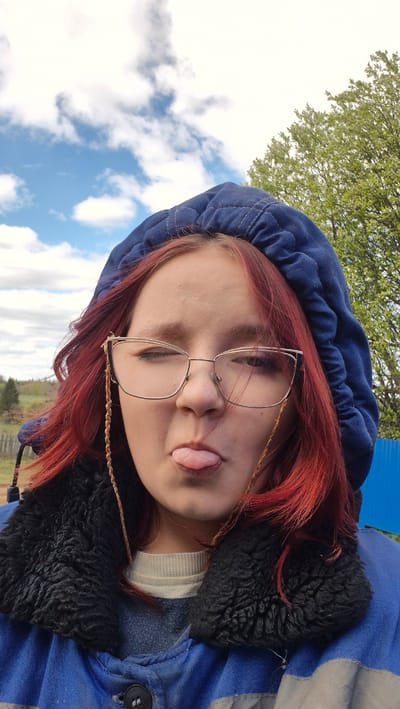 A person with red hair, wearing glasses and a blue hooded jacket with a black fur-lined collar, is sticking their tongue out. The background shows a partly cloudy sky, a tree with green leaves, and a blue structure. The image may have been taken outdoors in an area with some greenery. The location is Дойная, Russia.