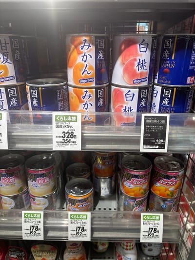 The image shows a refrigerated display case in a grocery store in Minato City, Japan. The case is stocked with canned fruit and fruit juice. There are a variety of brands, but all the cans feature Japanese writing. The cans are displayed in rows and are organized by type of fruit. There are price tags attached to some of the cans, indicating that the cans of fruit are sold for 328 yen and the cans of fruit juice are sold for 178 and 192 yen. The location of the grocery store is a commercial area in the Minato City, Japan.
