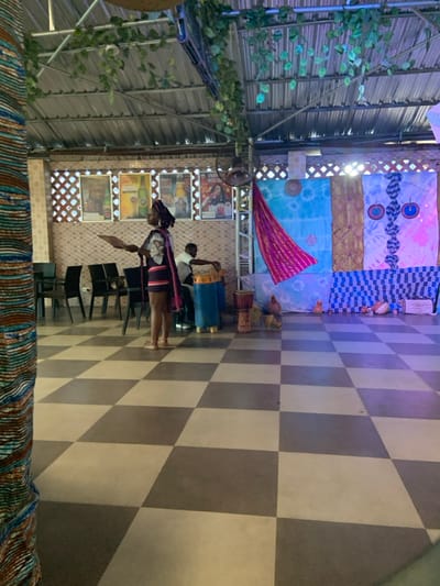 An indoor scene showcases a checkered floor. In the background, a wall is adorned with various patterned textiles and posters. A woman in traditional attire stands near a row of chairs. Drums and two chickens are positioned nearby. The ceiling structure is visible, with decorative foliage. The overall atmosphere appears vibrant and culturally rich.