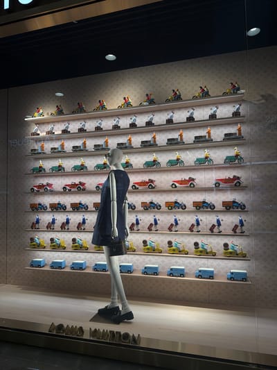 This is a photograph of a Louis Vuitton storefront display window in Boston, Massachusetts, USA. The display features multiple shelves filled with miniature decorative items. The background is a light-colored wall with a repeating Louis Vuitton logo pattern. On the shelves are rows of toy vehicles and figures, including yellow scooters with riders, blue vans, red cars, and figures dressed in blue pushing luggage carts. A mannequin dressed in a dark blue sleeveless dress and black shoes stands in the foreground, facing to the right, holding a small black handbag. The mannequin is positioned in front of the shelves, obscuring some of the display. The bottom of the window displays the "LOUIS VUITTON" logo in large metallic letters. The lighting appears to be artificial, casting shadows on the wall behind the display.