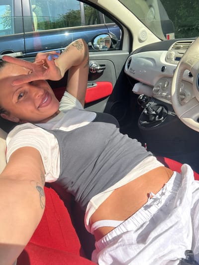 A bright, sunlit selfie shows a woman lying relaxed in the passenger seat of a car, likely a convertible given the strong overhead light. The image is oriented vertically with the woman's head towards the bottom right.
The woman is smiling at the camera, making a peace sign with her left hand, which has light blue nail polish. Tattoos are visible on both of her arms. She is wearing a grey ribbed tank top over a white top, with white drawstring shorts revealing a white undergarment strap.
The car's interior features a cream-colored dashboard with the text "500" clearly visible, identifying the vehicle as a Fiat 500. The car seats are a vibrant red. A black gear shifter in the center console has partially visible text that appears to say "aero". Reflections in the side mirror and window suggest an outdoor, sunny setting during the daytime. The overall impression is one of leisure and happiness.