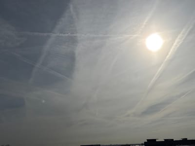 The image displays a bright, hazy sky over De Lier, Netherlands. A luminous, circular sun disc is prominently positioned in the upper right quadrant, casting a strong glare. The sky is permeated with numerous white contrails, appearing as linear and diffused streaks that crisscross each other. A distinct horizontal contrail spans the upper portion of the frame, intersecting with multiple diagonal contrails that extend across the sky. The horizon line, visible at the bottom, is dark and indistinct, showcasing the silhouettes of several low-lying structures or rooftops against the brighter sky. No people, specific actions, or interactions are present in the scene.