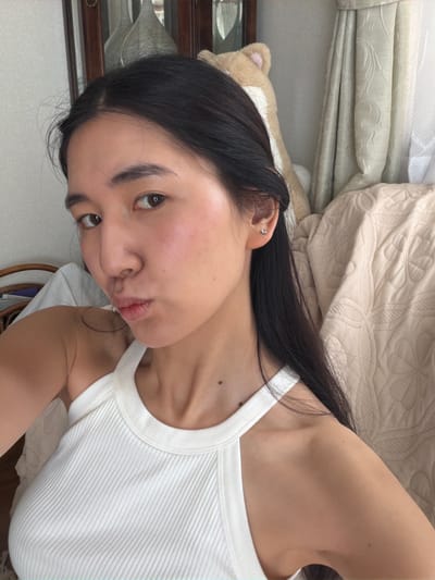 A young woman with long, dark hair is lying on a bed or similar surface covered with beige bedding. She is wearing a white ribbed tank top. She is facing the camera with a slight pout. A stuffed animal is visible near her head, along with what appears to be a wooden frame or piece of furniture. The image appears to have been taken indoors with a natural light source.

There is no indication of this picture being taken in Kawasaki, Japan.