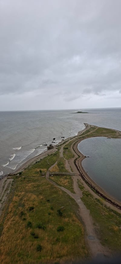 The aerial view showcases a narrow peninsula of land extending into a calm, grey body of water under an overcast sky. The peninsula is characterized by a strip of dry, yellow and green grass, with a winding dirt path visible along its length. Rocky outcrops and a few small structures are present along the water's edge on the left side of the peninsula. Further out in the water, a distant, narrow strip of green land can be seen. The right side of the peninsula is bordered by a larger, still body of water. The scene depicts a desolate natural landscape with minimal human presence, suggesting a remote coastal area. The location is identified as Sääre, Estonia.