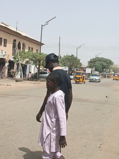 The image depicts an urban street scene in Hotoro South, Nigeria, under clear, bright daylight. In the immediate foreground, two individuals, both appearing to be male, walk away from the viewer on a gray asphalt road. The taller individual wears a dark t-shirt and a black baseball cap. The shorter individual, a child, walks slightly ahead and to the left, dressed in a light-colored, possibly pale lavender, long-sleeved tunic and trousers. Distinct shadows are cast on the sunlit pavement.
To the left of the road, a multi-story building with light-brown facades and arched windows stands, alongside street-level stalls displaying textiles and other goods. A discarded black tire rests near the curb. The street is lined with green, leafy trees and equipped with black streetlights featuring solar panels. Overhead utility wires are visible.
The mid-ground and background reveal an active urban thoroughfare. Multiple yellow motorized tricycles (keke napep) are present, some moving, some potentially parked. A silver sedan drives towards the viewer in the middle distance. Other vehicles and indistinct pedestrians are visible further down the road, contributing to the busy street environment.