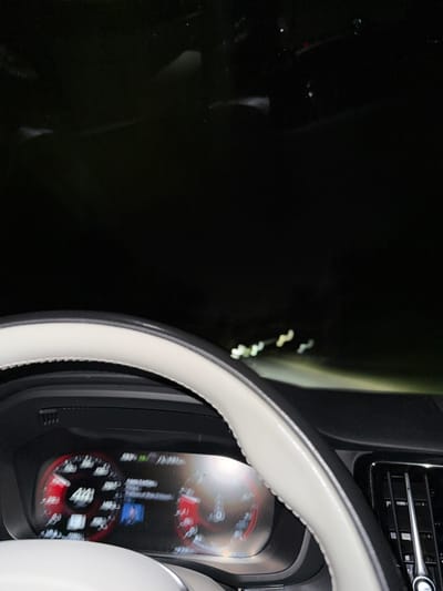 The image presents a nighttime view from the driver's perspective inside a vehicle. A light-colored steering wheel with dark trim occupies the lower-left and central foreground. Behind it, a digital instrument cluster is illuminated, displaying a speed of "44" (speed unit not explicitly visible) on the left side, alongside other vehicle metrics and a central multimedia display showing a song title and a small image. A dark dashboard is visible, with a vertical air vent on the far right. The vehicle's headlights illuminate a dark road ahead, and a series of blurry, distant white lights are visible in the upper-right portion of the exterior view. No individuals are visible. The environment appears dark and sparsely lit, consistent with night driving on a road outside a brightly lit urban area. The precise location within West Windsor, United States, cannot be determined from the visual information provided in the image.