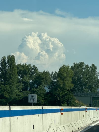 This image was taken on the E45 highway near Città di Castello, Italy. The scene is dominated by a large, fluffy cumulus cloud in a bright blue sky. Below the clouds, a dense line of green trees stretches across the middle ground. In the foreground, a concrete barrier with a bright blue stripe runs along the edge of the highway. A white sign with the Roman numeral "IV" and the number "116" is visible on a post in front of the trees. The perspective suggests the photo was taken from a moving vehicle.