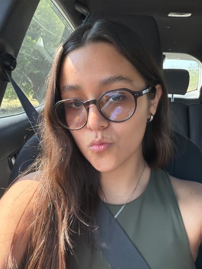 A young woman wearing glasses and a green top is reclining in a car seat. She has dark hair with warm highlights, a silver necklace, and a ring on her left ear. The sunlight is illuminating her hair and face, creating a soft, diffused light. A car window is visible in the background, and a blurred landscape is visible through the glass.