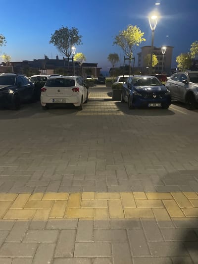 The image depicts a parking lot at dusk in Yunus Emre, Turkey, likely near a commercial area given the buildings in the background and the presence of streetlights. Several cars are parked, including a white SEAT Ibiza with license plate "43 ACF 09B" in the foreground, a dark blue car with license plate "45 AUN 148", and a silver car to its right. A dark-colored car is partially visible on the left. The parking lot is paved with interlocking bricks, with a distinct yellow brick pattern running horizontally in the foreground. Modern streetlights with illuminated, curved fixtures are spaced throughout the area, casting a warm glow on the scene. Trees with illuminated yellow foliage are planted near the streetlights. The sky is a deep twilight blue, indicating the transition from day to night. The overall environment suggests an urban or suburban setting during evening hours.