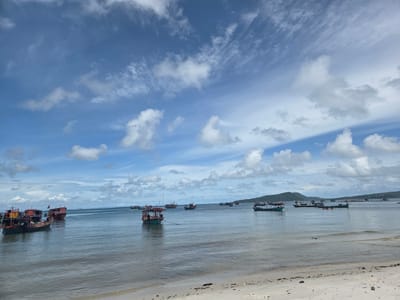 The image depicts a serene coastal scene in Koh Toch Village, Cambodia, under a bright, cloudy blue sky. The foreground features a sandy beach with scattered debris, leading to calm, shallow ocean waters. A fleet of small, colorful fishing boats is anchored in the middle ground, some appearing to be wooden with red and green accents. The boats are clustered together, indicating a harbor or a resting area for the vessels. In the background, lush green hills rise from the coastline, suggesting a tropical island setting. The overall atmosphere is tranquil and depicts a typical day in a coastal fishing community.