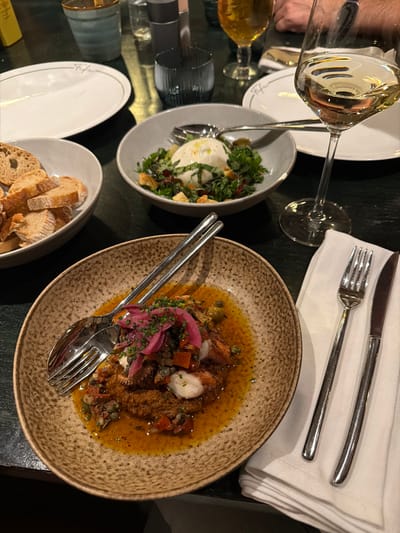 A table setting displays a meal with multiple courses. There's a plate of sliced bread, a bowl of burrata salad with greens, and a plate containing octopus with a reddish sauce, capers, and pickled onions. Flatware is arranged near each dish, along with a folded napkin and a knife. Two wine glasses, one filled with a clear liquid and the other with a darker liquid, are visible. The overall presentation suggests a formal or upscale dining experience.