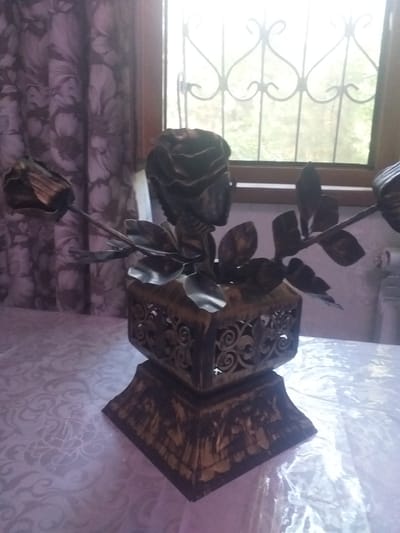 The image displays a wrought iron sculpture of a rose with several leaves, resting on a decorative, possibly wooden, box with intricate scrollwork. The sculpture is dark metal, likely iron, with a patina that suggests age or a specific finishing technique. One rose bud is prominently featured in the foreground, with another bud and more leaves trailing upwards. The box the sculpture is placed on is ornate, with a pattern of swirling designs and possibly carved or painted imagery on its sides.

The setting appears to be indoors, near a window. Through the window, we can see greenery and what looks like a decorative metal grate or window bars. The light coming from the window suggests it is daytime. The background includes a patterned curtain or fabric, adding a domestic or decorative element to the scene. The overall impression is one of an artistic display, possibly in a home or gallery setting in Navoiy City, Uzbekistan. There is no visible text in the image. The lighting and composition of the image are not ideal, with some blurriness and overexposure from the window light, making it difficult to discern finer details.
