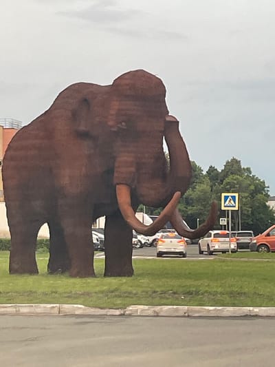 A large, reddish-brown mammoth sculpture stands prominently on a grassy area, facing to the right. The sculpture is detailed, showing tusks, ears, and a trunk. Behind the sculpture, a road is visible with several white cars driving and parked. A pedestrian crossing sign with a blue background and white pedestrian symbol is present to the right of the mammoth. Trees form a backdrop behind the road and cars. In the far left background, a multi-story building with a light-colored facade and railings is partially visible. The sky is overcast and light gray. This scene is located in the city of Novoye Shigaleevo, Russia.