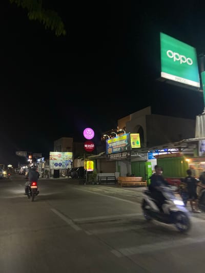 This is a nighttime street scene in the city of Crewek, Indonesia. A road runs through the center of the frame, with businesses and illuminated signs lining both sides. On the left, a motorcycle with a rider is moving away from the camera, its taillight illuminated. Further down the road, another motorcycle is approaching, its headlight on.

Several businesses are visible. A large green sign with the word "OPPO" in white lettering is prominently displayed high on the right. Below it, a smaller sign with "PIRIT CELL" is visible. On the left, circular signs for "all of Aesthetic" and "waslè Cafe & Resto" are lit in pink and red, respectively. A large banner with colorful food images and text in Indonesian, along with smaller signs advertising phone services, is also present. Further down the street, a bright yellow sign reads "SERVICE HP" with an Apple logo.

The environment appears to be urban, with buildings constructed from various materials and street lighting casting shadows. The road surface is asphalt. The overall atmosphere is that of a commercial street at night.