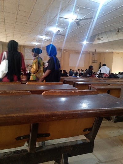This image captures the interior of a large lecture hall or classroom in Alabata, Nigeria. The scene is dominated by multiple rows of fixed, dark brown wooden desks with attached seats, supported by dark metal frames, extending from the foreground into the background. The ceiling features off-white tiles, several dark-bladed ceiling fans, and bright fluorescent light fixtures producing prominent lens flares. The walls are light brown, with rectangular white boards visible on the back wall.

In the mid-ground, three individuals are positioned between desk rows. From left to right: a person with long dark hair, wearing a dark garment and carrying a white shoulder bag; a person in a patterned yellow, brown, and green long-sleeved top with a dark head covering; and a person wearing a blue head covering, a dark t-shirt with a white graphic, and carrying a dark bag. Their backs are generally towards the camera or they are in profile. In the background, numerous additional individuals are seated at desks, partially obscured by the foreground elements. One individual on the far right in the background has a white cloth draped over their shoulders. The environment indicates a communal learning space.