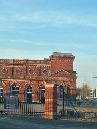 The image displays the Copper Rivet Distillery and Pumproom building, located on Leviathan Way in Chatham, United Kingdom. The prominent structure is a multi-story, red brick building characterized by decorative yellow brick banding, arched windows on the lower level, and circular windows on the upper level. A distinctive square tower section, topped with a weathered grey cap, is visible on the building's right side. The sky above is clear blue with sparse, wispy white clouds. In the foreground, a dark metal fence with vertical bars runs across the scene, separating an asphalt surface from the building. A brick pillar matching the building's facade stands in the mid-foreground. A black sign affixed to the fence, partially obscured, states "WELCOME TO COPPER RIVET DISTILLERY and PUMPROOM". To the right of the brick pillar and behind the fence, an open area features concrete blocks, chain-link fencing, and in the distant background, other buildings and bare trees. No individuals are present in the scene.