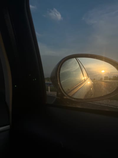 The image is a close-up shot of a car's side mirror, reflecting a road scene at sunset. The mirror is oval-shaped and mounted on the exterior of a vehicle. The reflection shows a highway stretching into the distance, with the sun low on the horizon, casting a warm golden light. The sky is a mix of blue and orange hues, with scattered clouds. The reflection also captures part of the car the photo is taken from, specifically the rear passenger door and window frame. The foreground is dominated by the interior of the car, with the dark trim of the door and window. In the background, outside the mirror's reflection, is a glimpse of a blue sky with white clouds. There are no people or animals visible in the image. The location in the city of Saraia, Bulgaria is not identifiable from the image.