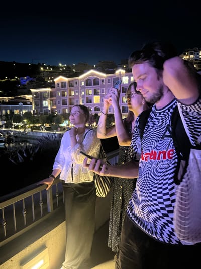 Three individuals are standing on a balcony at night, overlooking a well-lit town situated on a hillside. The town appears to be comprised of multi-story buildings with illuminated windows and decorative exterior lighting. In the foreground, a man is captured from the chest up, with his hand running through his hair. He is wearing a black and white patterned t-shirt with red lettering. To his left, a woman with glasses is holding a smartphone, seemingly taking a photo or video. Further to the left, another woman, seen from the chest down, is wearing a white, eyelet-embroidered top and a long dark skirt. She is leaning on a railing, holding a smartphone in her hand. The balcony has a metal railing and a concrete wall below. The overall scene suggests a view of the coastal town of Gošići, Montenegro, at night.