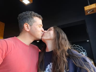 The image shows a couple kissing. The woman has long, brown hair and is wearing a dark-colored shirt with a white design. The man has short, grey hair and is wearing a red shirt. The background is dark, and there are a few objects visible, including a small wooden box with something white inside. The photograph appears to be rotated. There is no evidence to suggest the image was taken in Sarandí, Argentina.