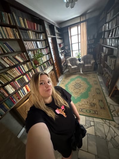 A woman with blonde hair is taking a selfie in a room with floor-to-ceiling bookshelves filled with books. She is wearing a black t-shirt with a cartoon character on it and black shorts. In the background, there are two armchairs, a window with curtains, and a patterned rug on the floor. The room appears to be a library or study. The image is taken from a slightly high angle, looking down on the woman and the room. The lighting is dim, with light coming from the window. The woman is looking at the camera and has her mouth slightly open.