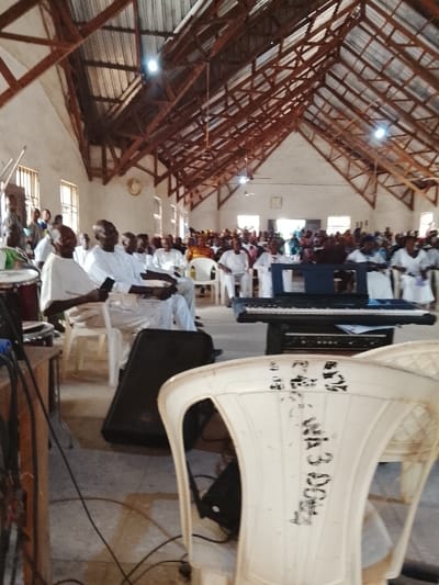 The image shows the interior of a building with a wooden truss roof and white walls. There are numerous people sitting in white plastic chairs, with a few standing. The people are mostly dressed in white clothing and are facing the front of the room. At the front, a black electronic keyboard is on a black stand. The camera is focused on the back of a white plastic chair with black markings. There is a black speaker on the floor next to it. The location is an open space with a tiled floor. The building appears to be a community center or a church, and the scene could be part of a religious service or a gathering of people. The coordinates provided (latitude: 8.4, longitude: 8.342) are near the city of Mukaiya, Nigeria.