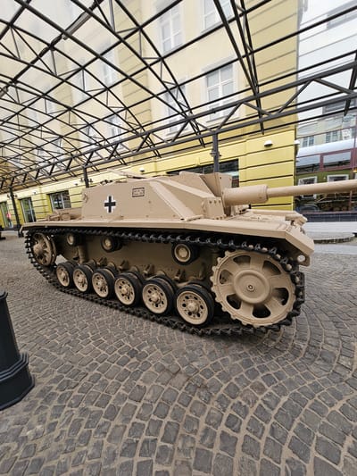 The image depicts a Panzerjäger 38(t) tank destroyer, likely a replica or restored vehicle, situated on a cobblestone surface. The tank is beige and features a black cross insignia. The scene appears to be an outdoor display, possibly within a museum or historical park. In the background, there are buildings reflecting in a glass ceiling with a metal frame. This aligns with the location Архангельское, Russia, which is known for its historical estate museum complex. The focus is on the tank itself, highlighting its features and placement within the environment.