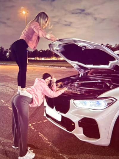 Two young women are interacting with the open hood of a white car at night, possibly in a parking lot. The woman on the left, wearing a pink jacket and black leggings, is standing on the rear of the woman on the right. The woman on the right, wearing a pink hoodie and grey pants, is leaning over the car's engine. The car's headlights are on, illuminating the engine compartment and the immediate surroundings. The background suggests an outdoor urban setting with a street light visible, and trees or buildings in the distance. The sky is dark with some clouds. The scene is shot from a slightly low angle.